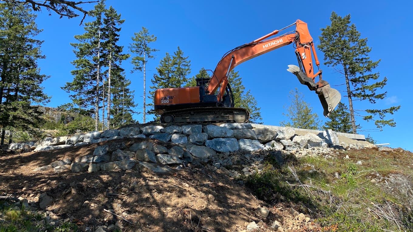 A Charlie's Excavating machine standing on a finished armour-rock retaining wall on Salt Spring Island, against a clear blue sky