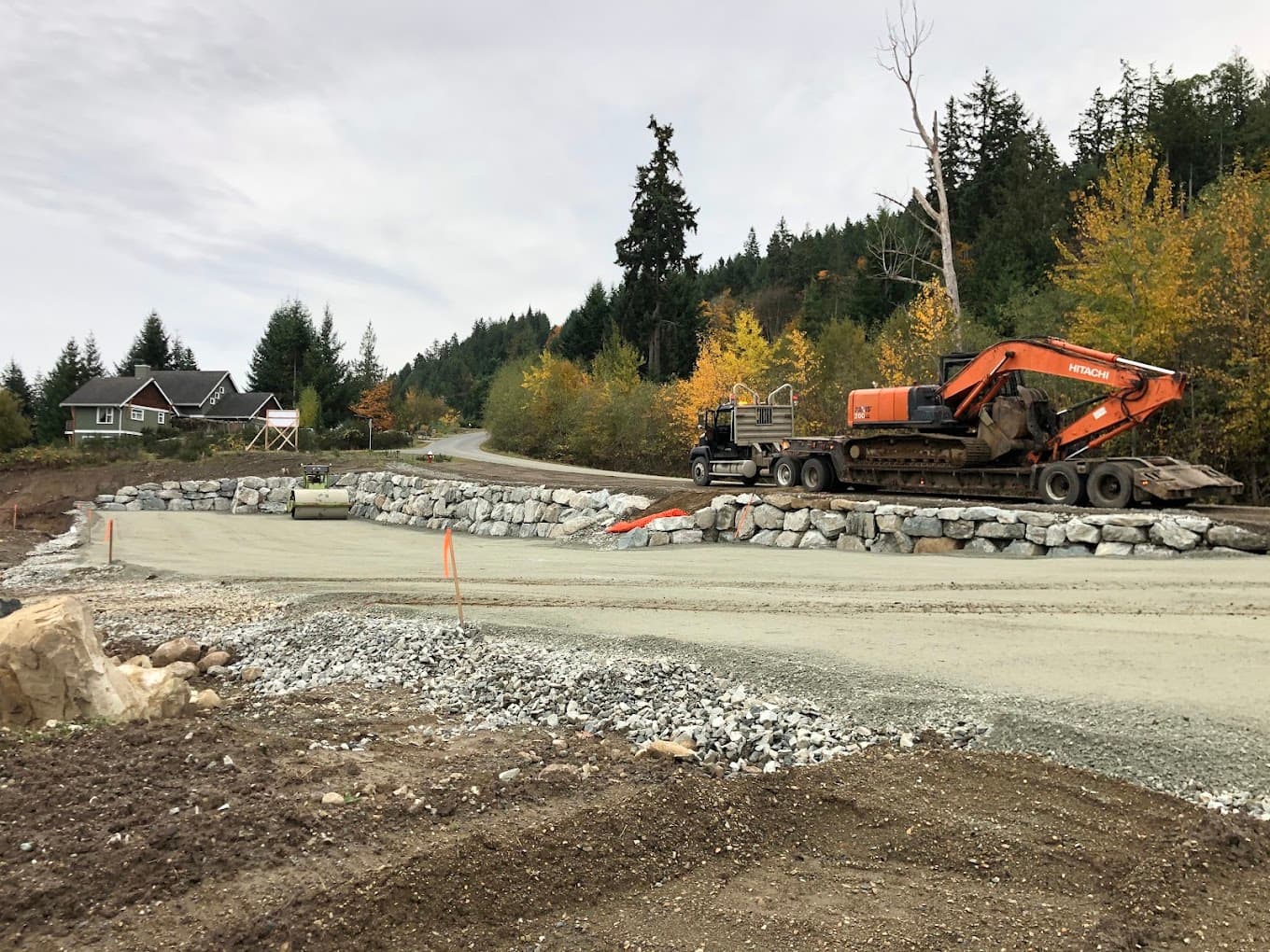 A low-bed truck loaded with an excavator beside a finished rock wall and gravel pad